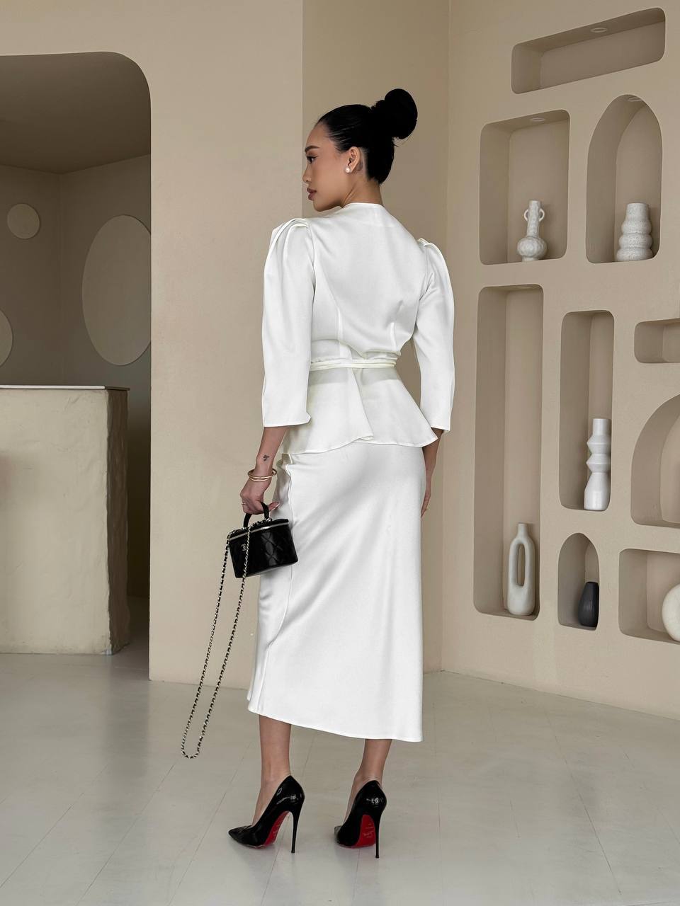 Woman in a white outfit holding a black handbag in a minimalistic room.