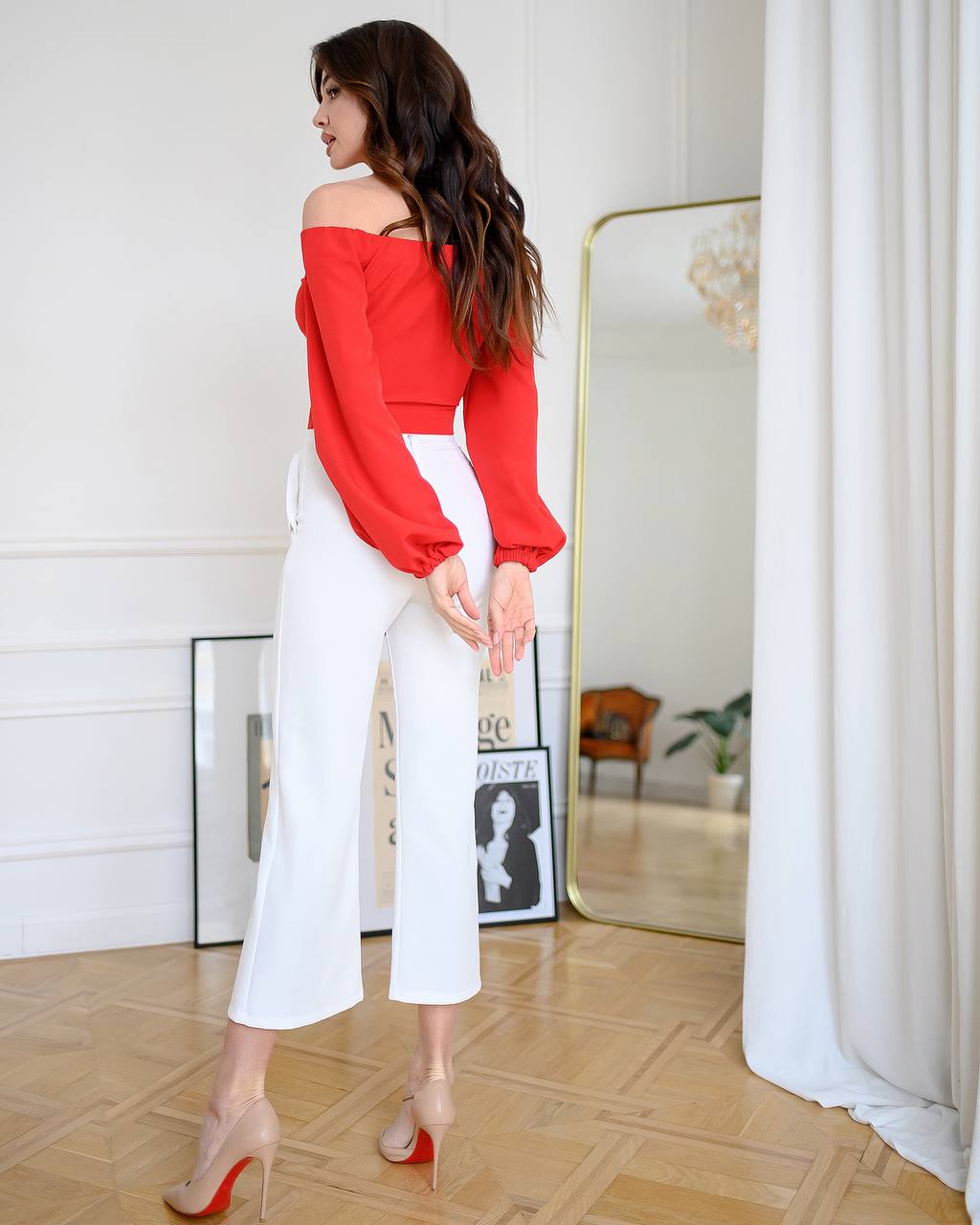 Woman wearing a red off-shoulder top and white pants in a room with a mirror and decor.