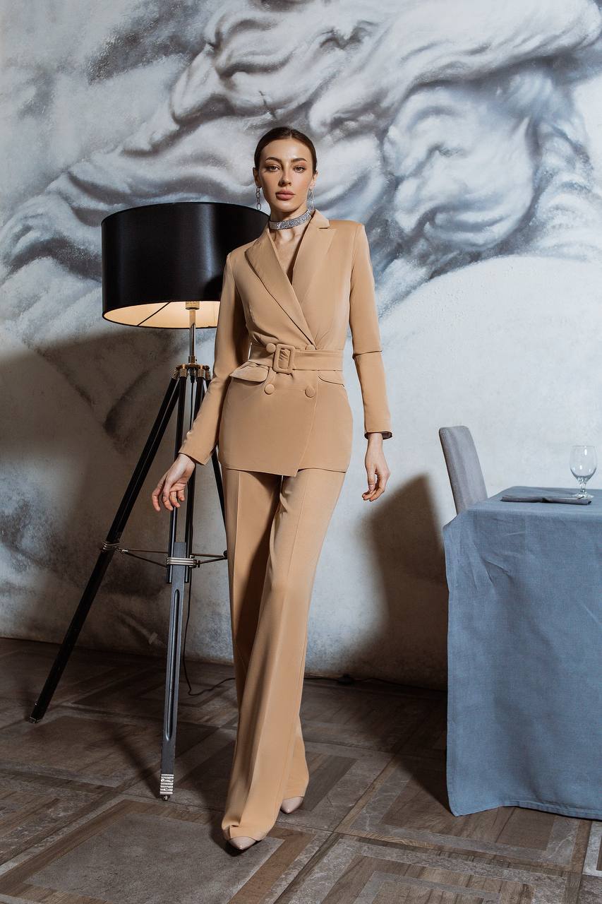 Woman in a beige suit standing in a room with a lamp and table.