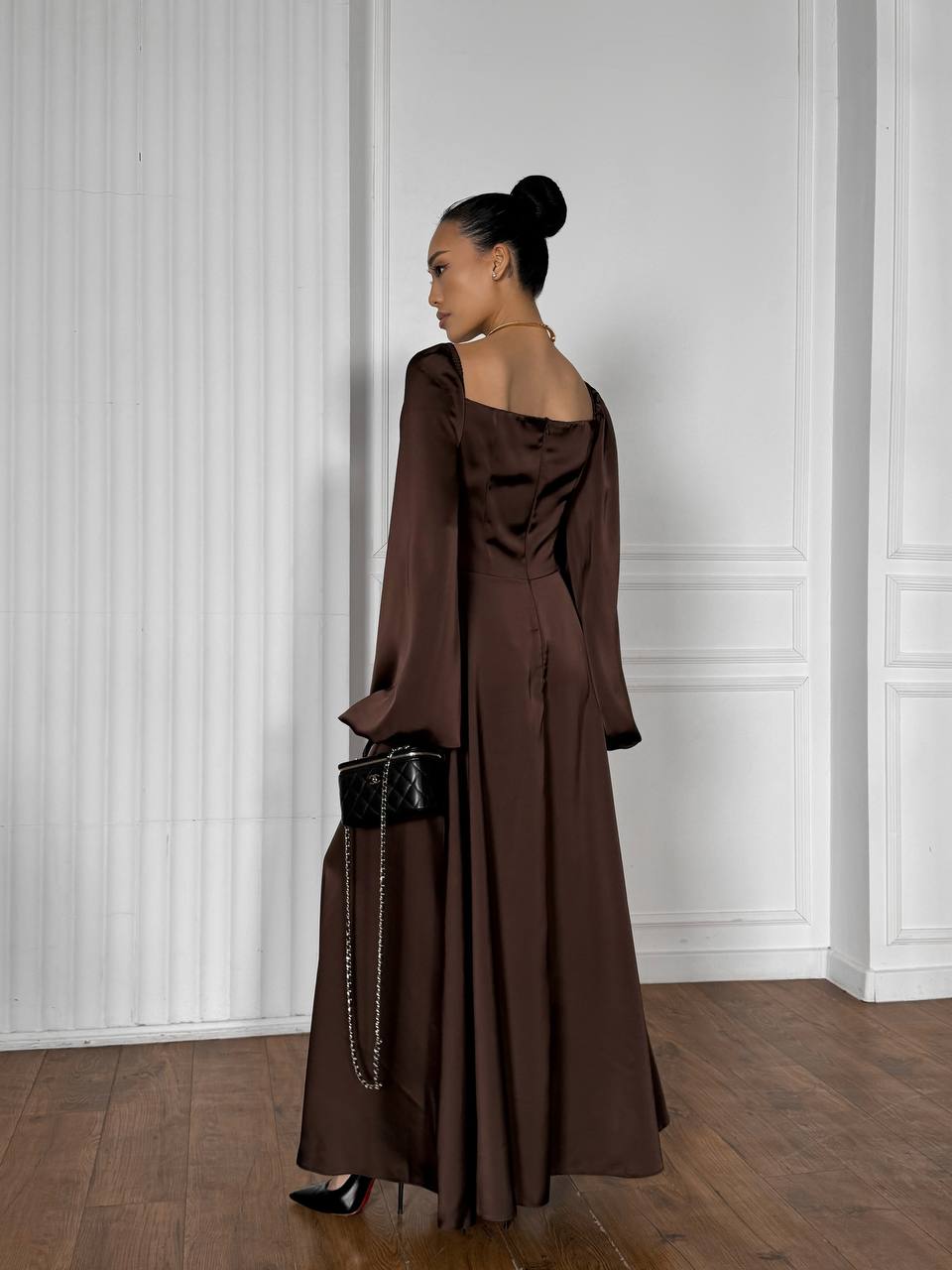 Woman in a brown off-shoulder dress standing against a white paneled wall.