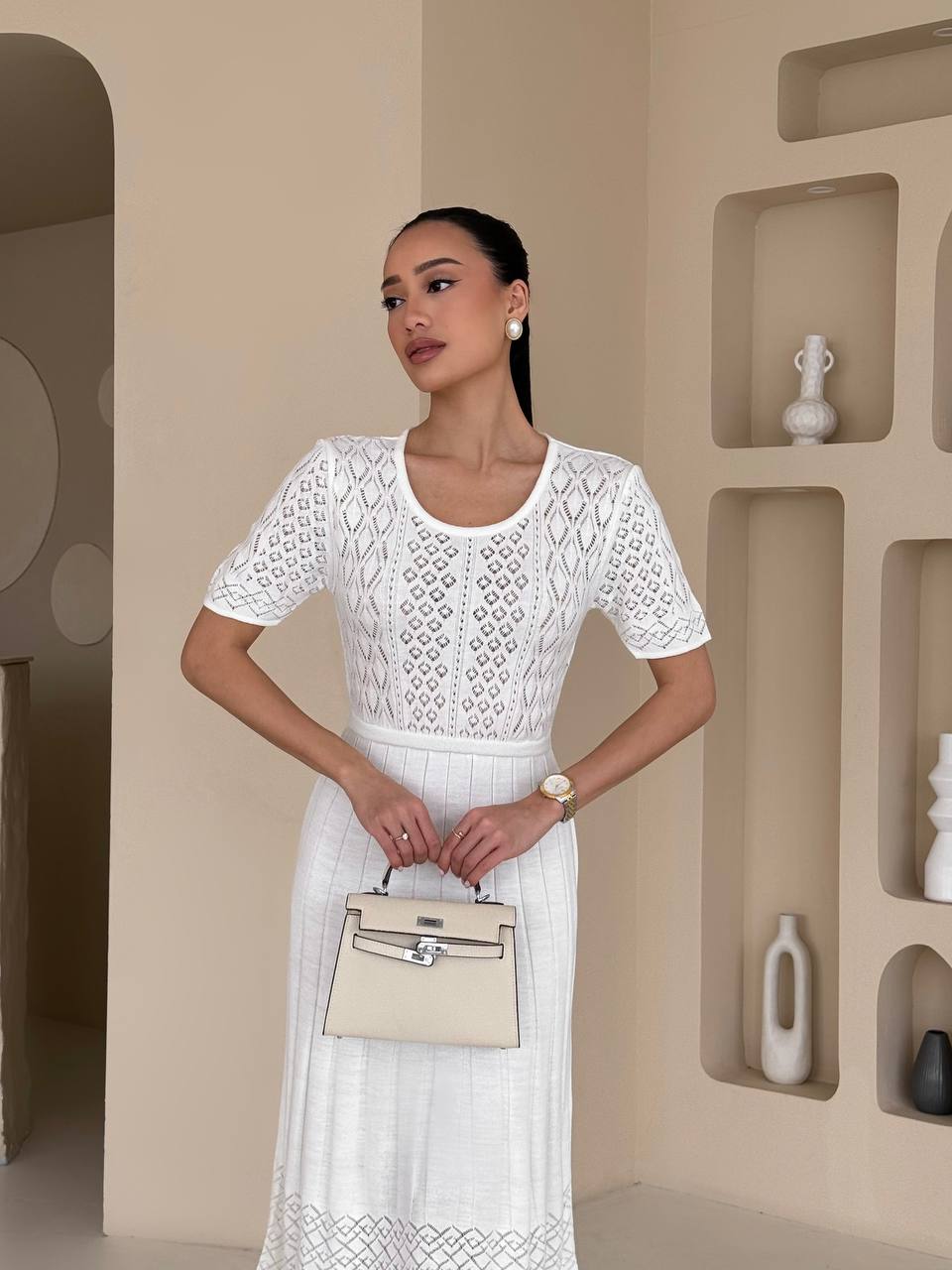 Woman in a white lace dress holding a beige handbag in a neutral setting