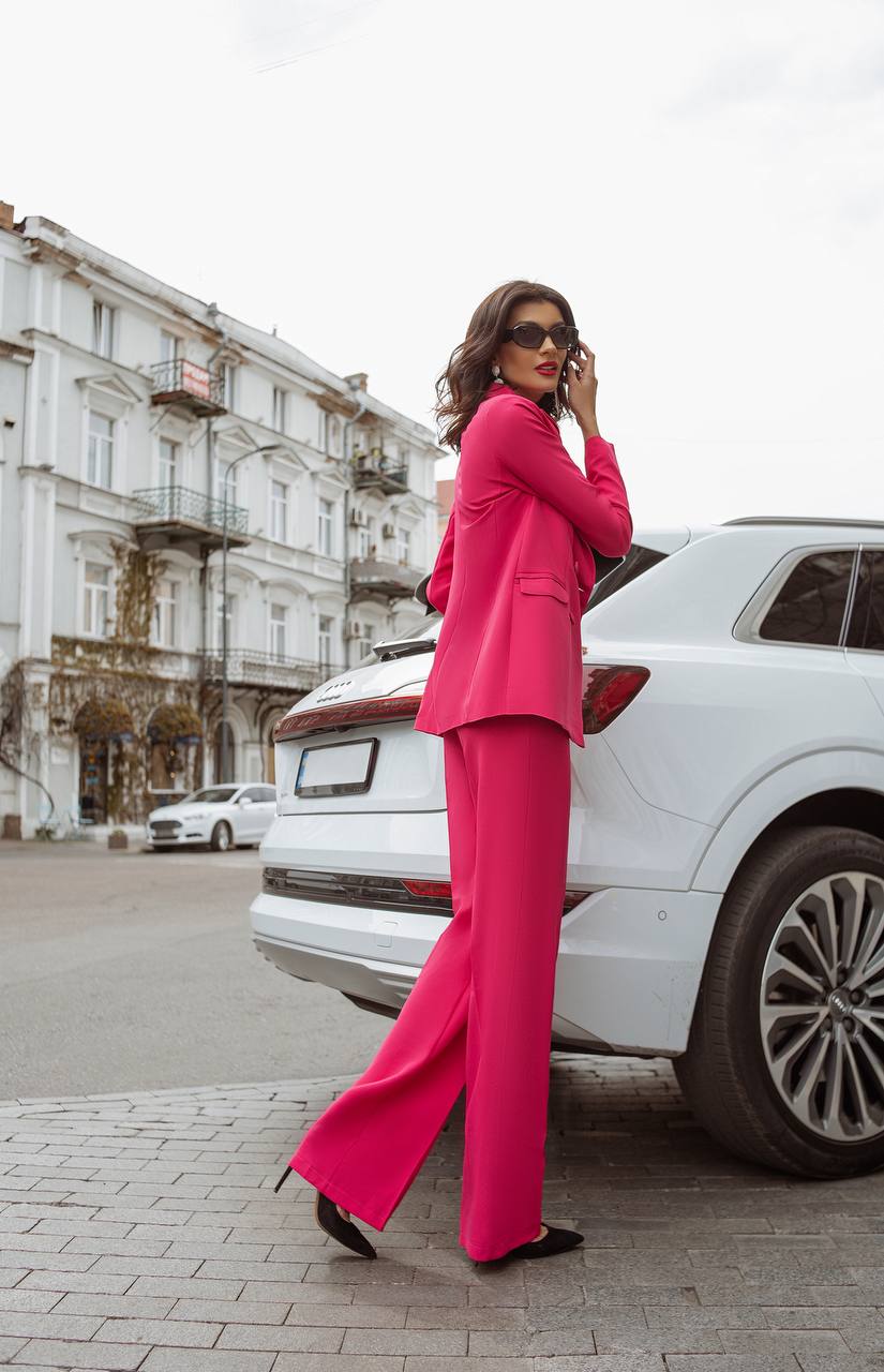 Woman in a bright pink suit standing on a city street with a white car and building in the background.
