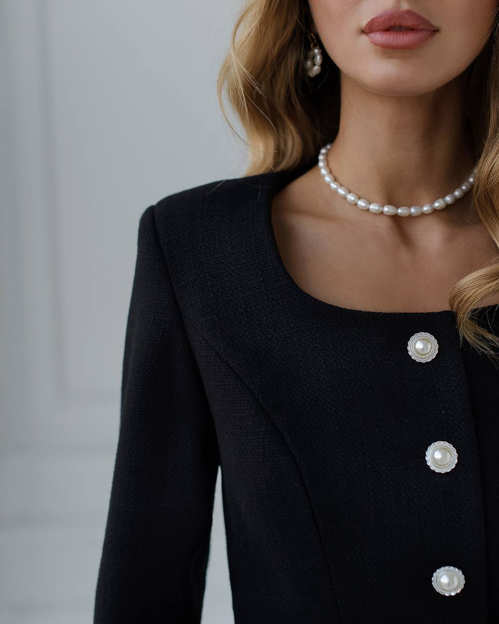 Close-up of a person wearing a black blazer with pearl buttons and a pearl necklace against a neutral background.