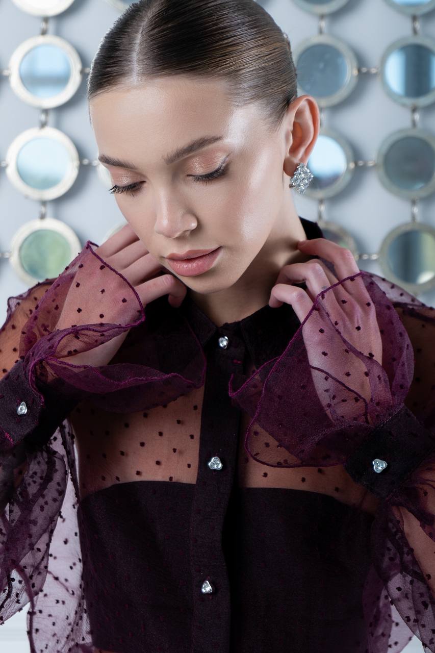 Woman wearing a dark blouse with sheer purple sleeves featuring small embellishments against a decorative wall.