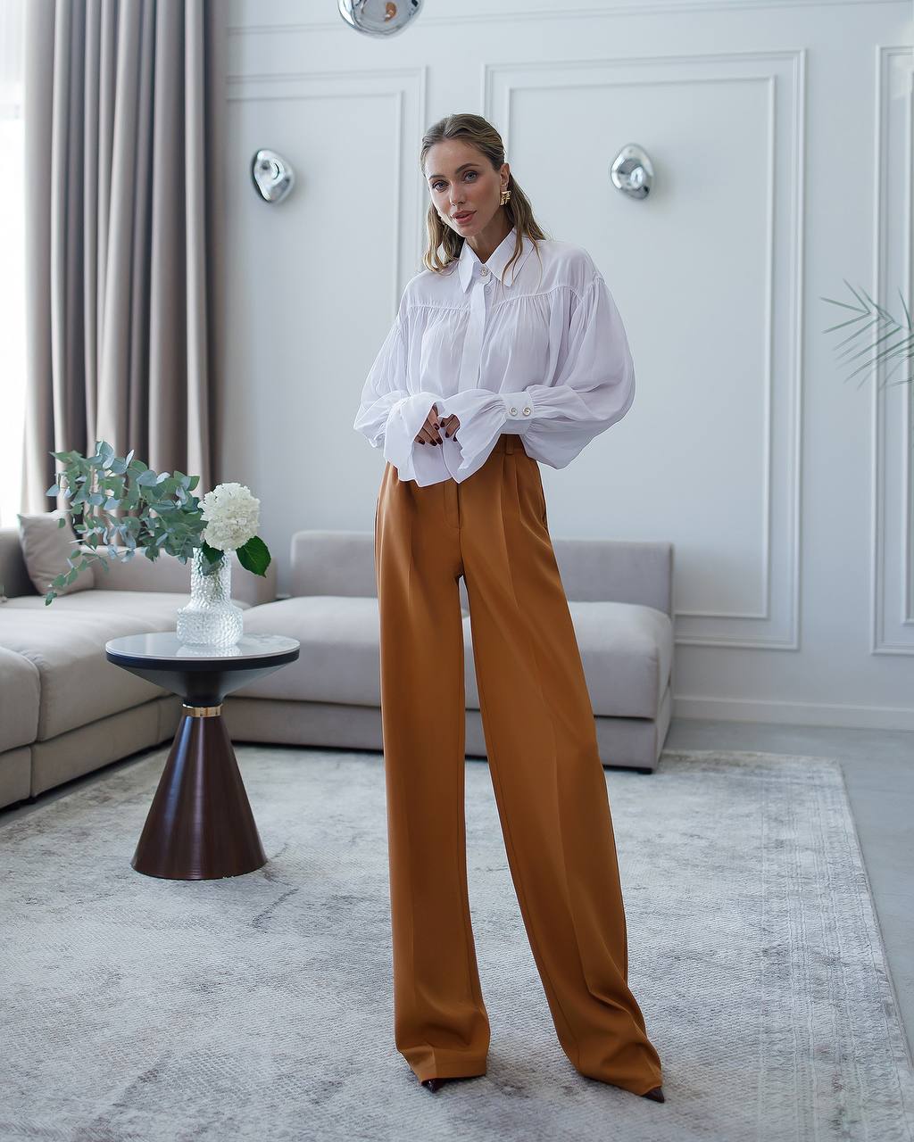 Woman in a white blouse and brown pants standing in a modern living room.
