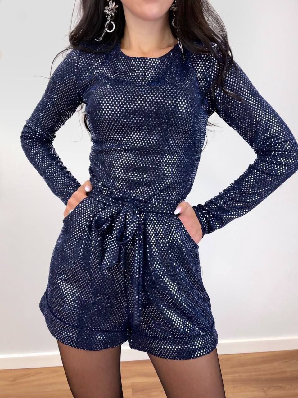 Person wearing a blue sequin outfit with long sleeves and shorts against a white wall.