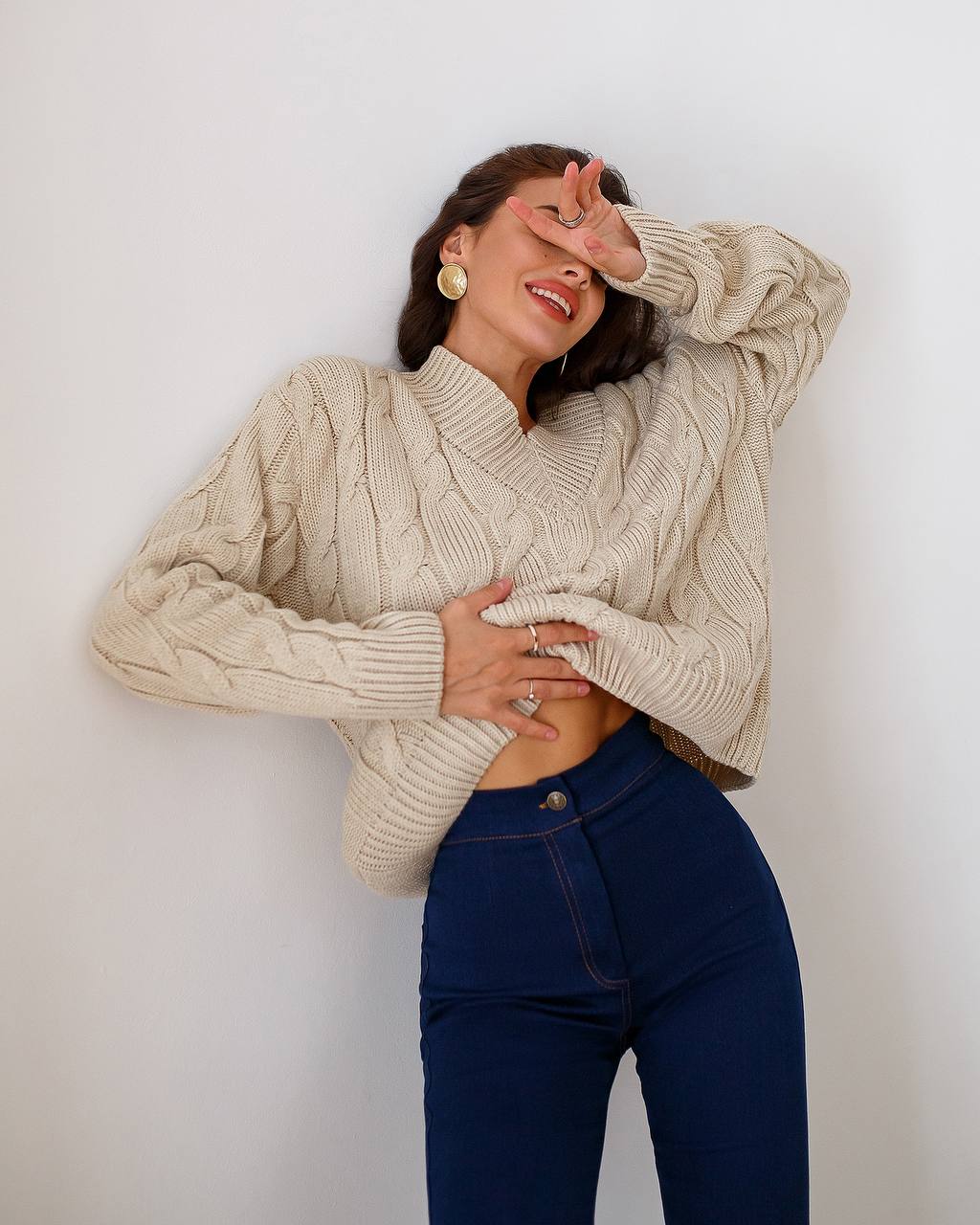 Woman wearing a beige cable knit sweater and blue jeans against a white background