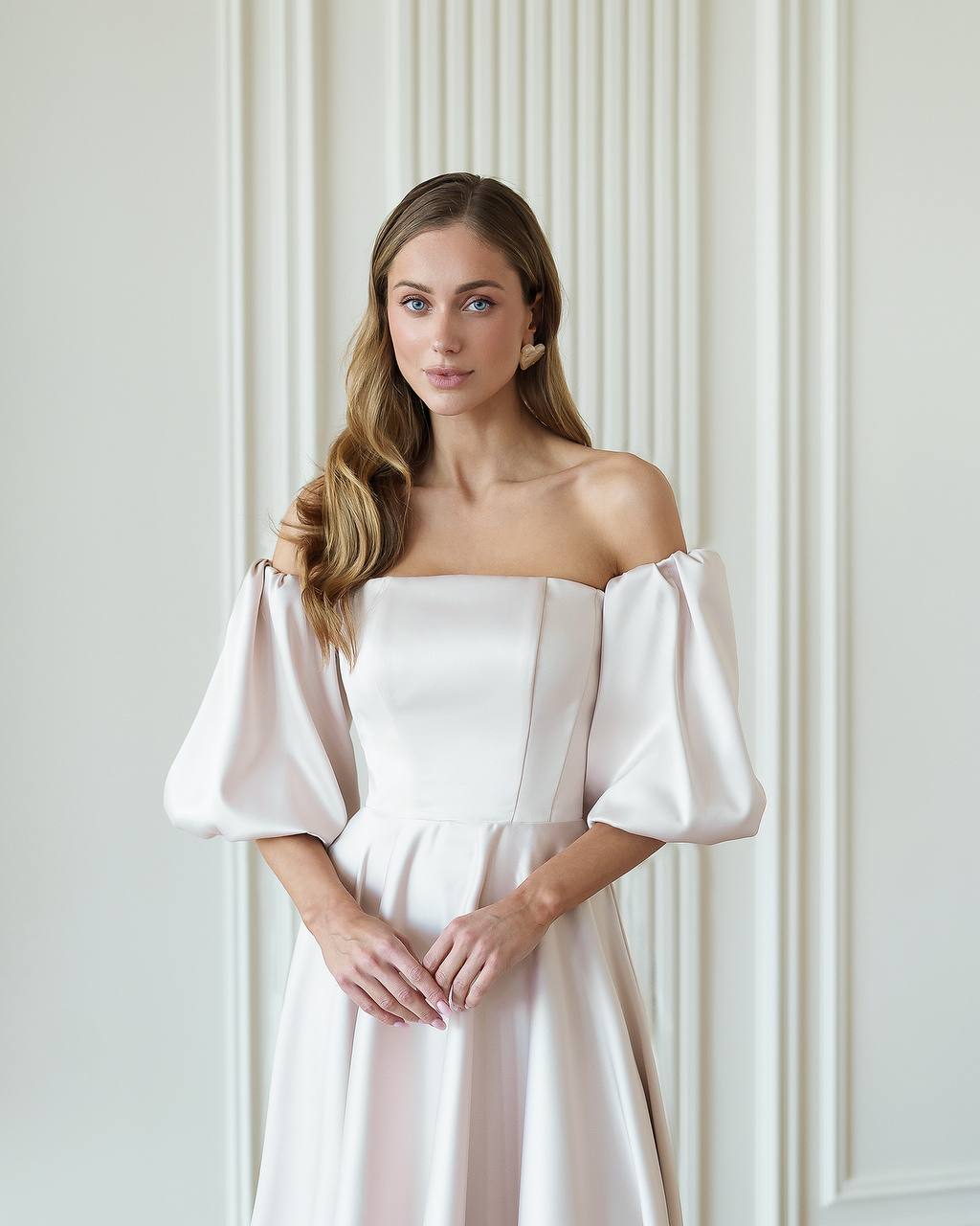 Woman wearing a white off-shoulder dress against a neutral wall.