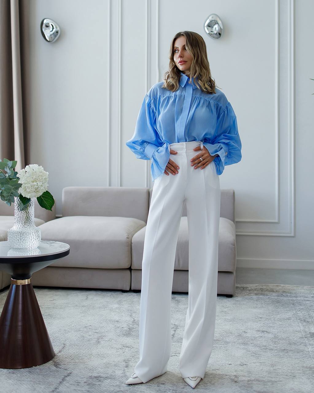 Woman in a blue blouse and white pants standing in a stylish room with a sofa and decorative items.