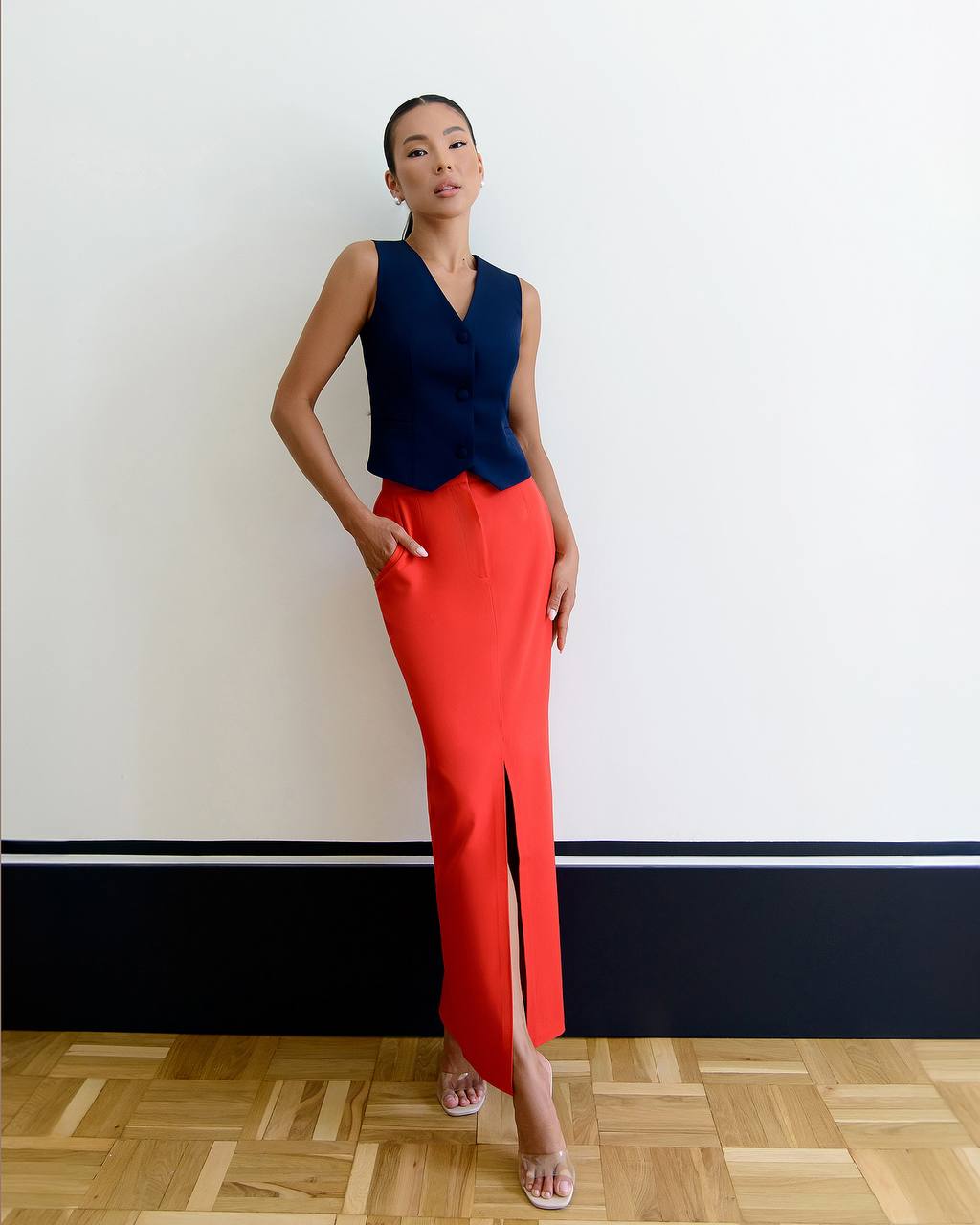 Woman wearing a navy top and red pants standing against a white wall.