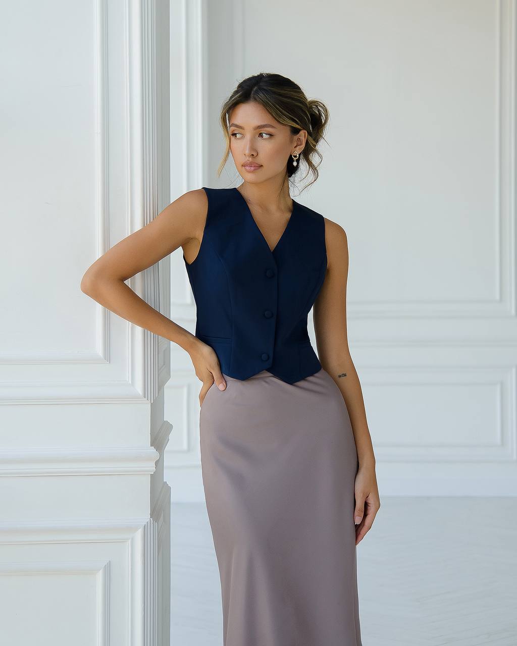 Woman wearing a navy vest and long beige skirt against a white wall.