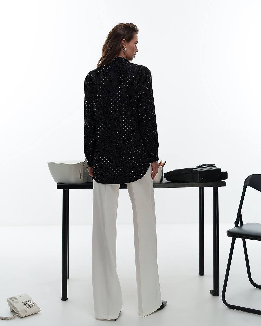 Woman standing in a minimalistic room with a black polka dot shirt and white pants.