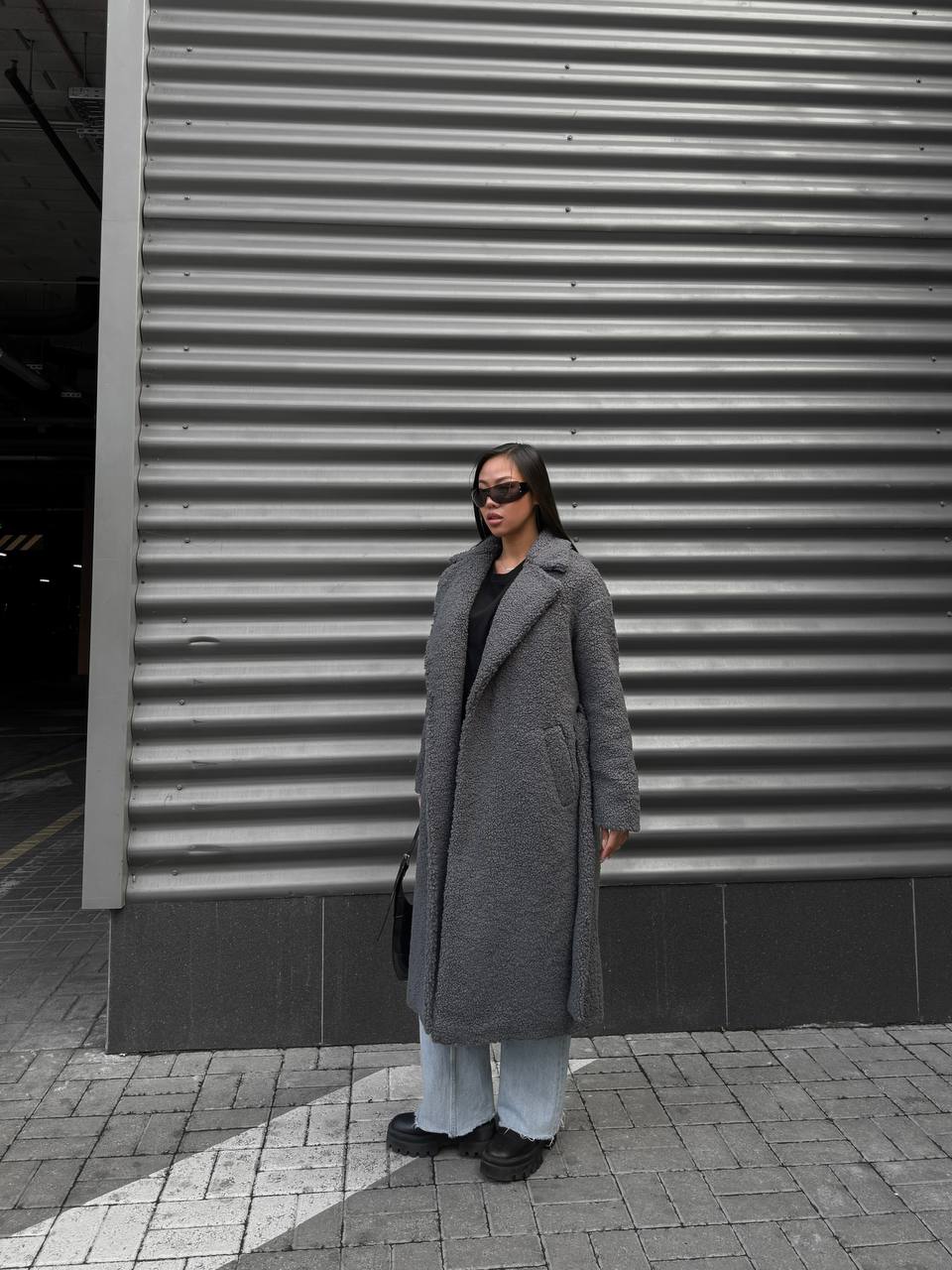 Person wearing a long gray coat standing in front of a corrugated metal wall.