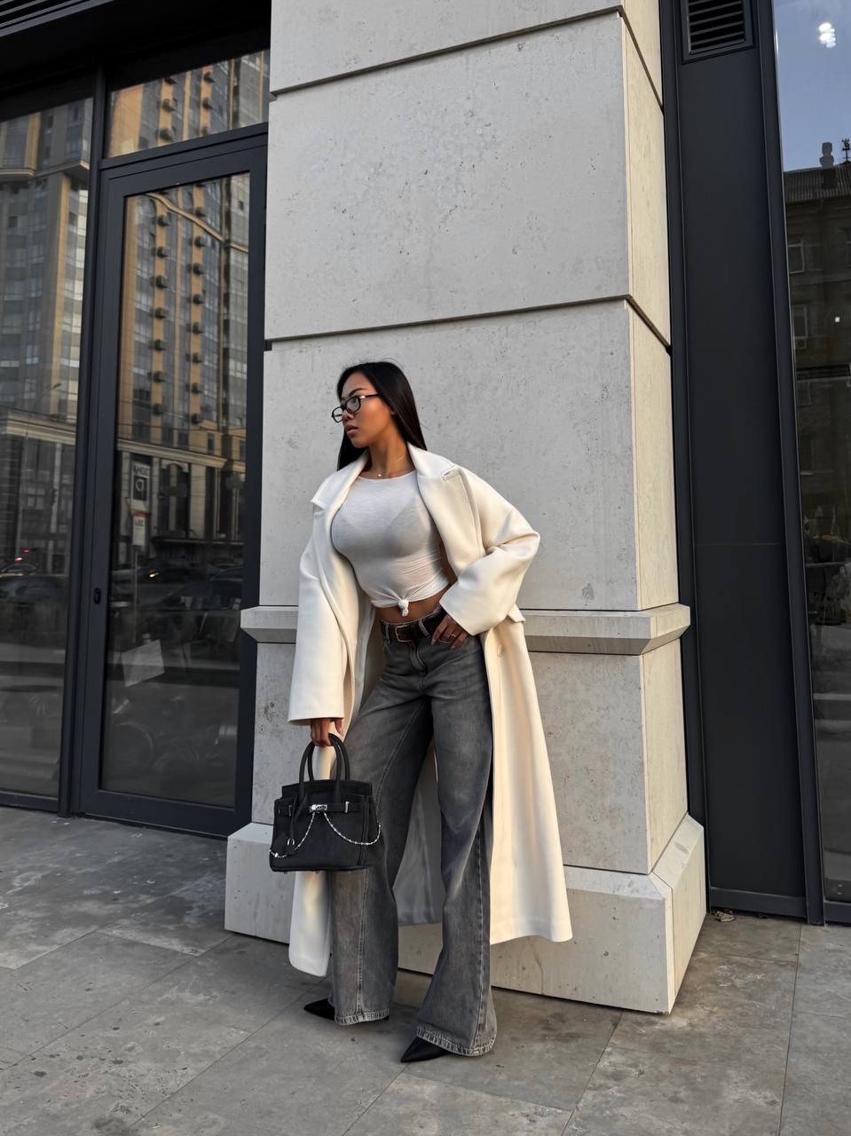 Woman in a white coat and gray pants standing against a building.