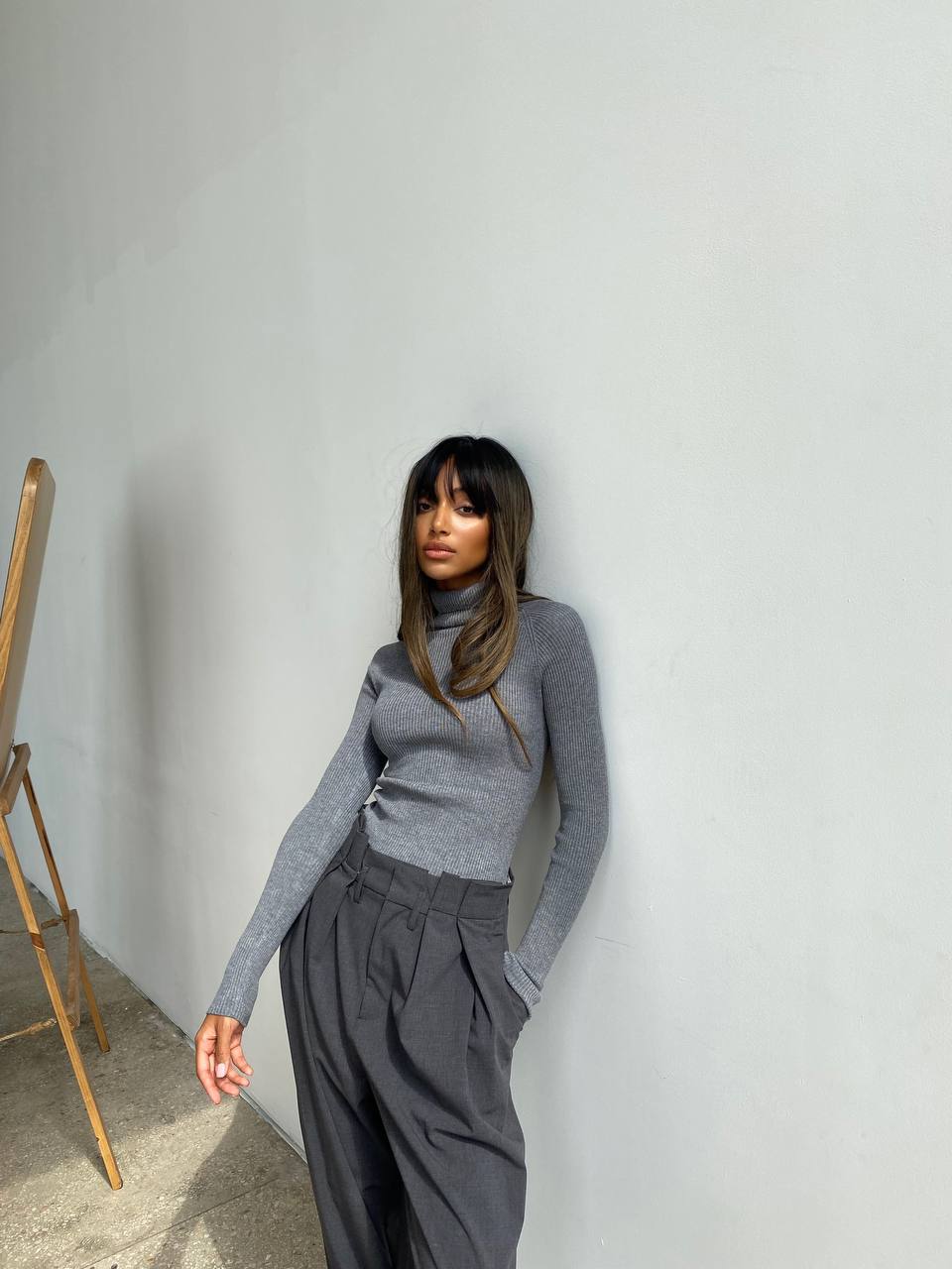 Woman wearing a gray turtleneck and wide-leg pants against a white wall.