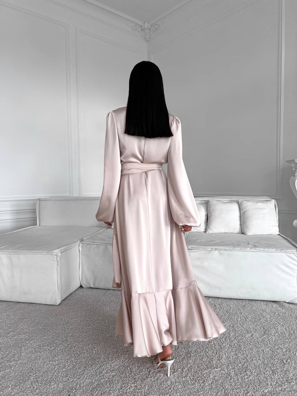 Woman in a long, light pink satin dress standing in a room with white walls and furniture.