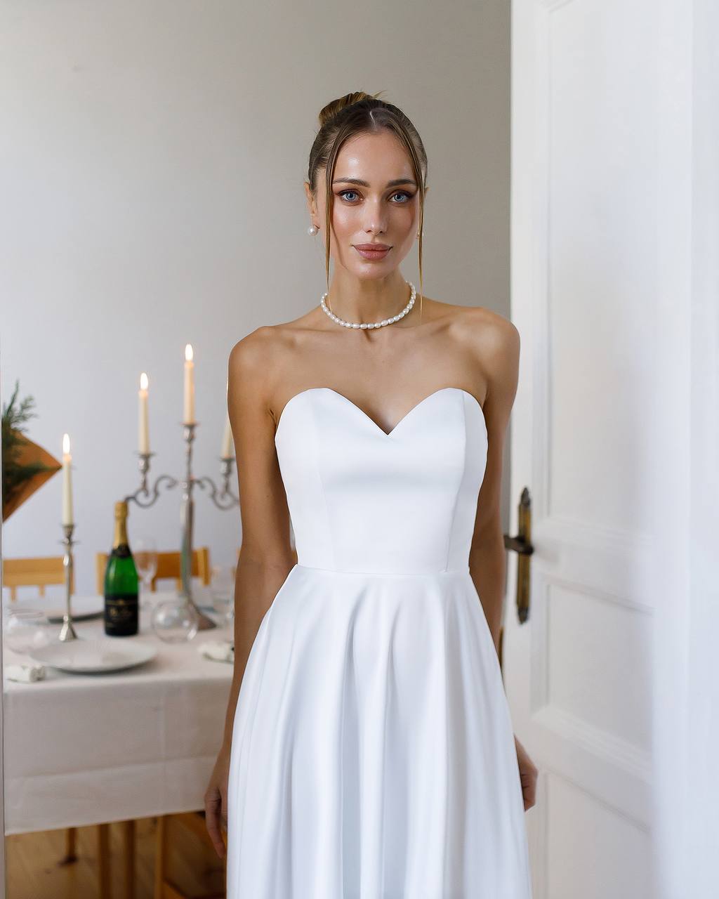 Woman in a white strapless dress standing in a room with a dining table and candles.