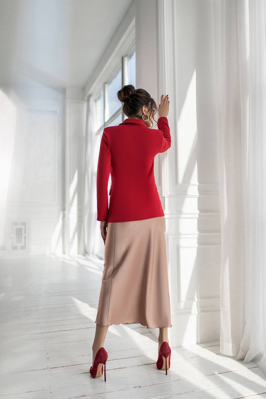 Woman in a red sweater and beige skirt standing in a bright room with white curtains.