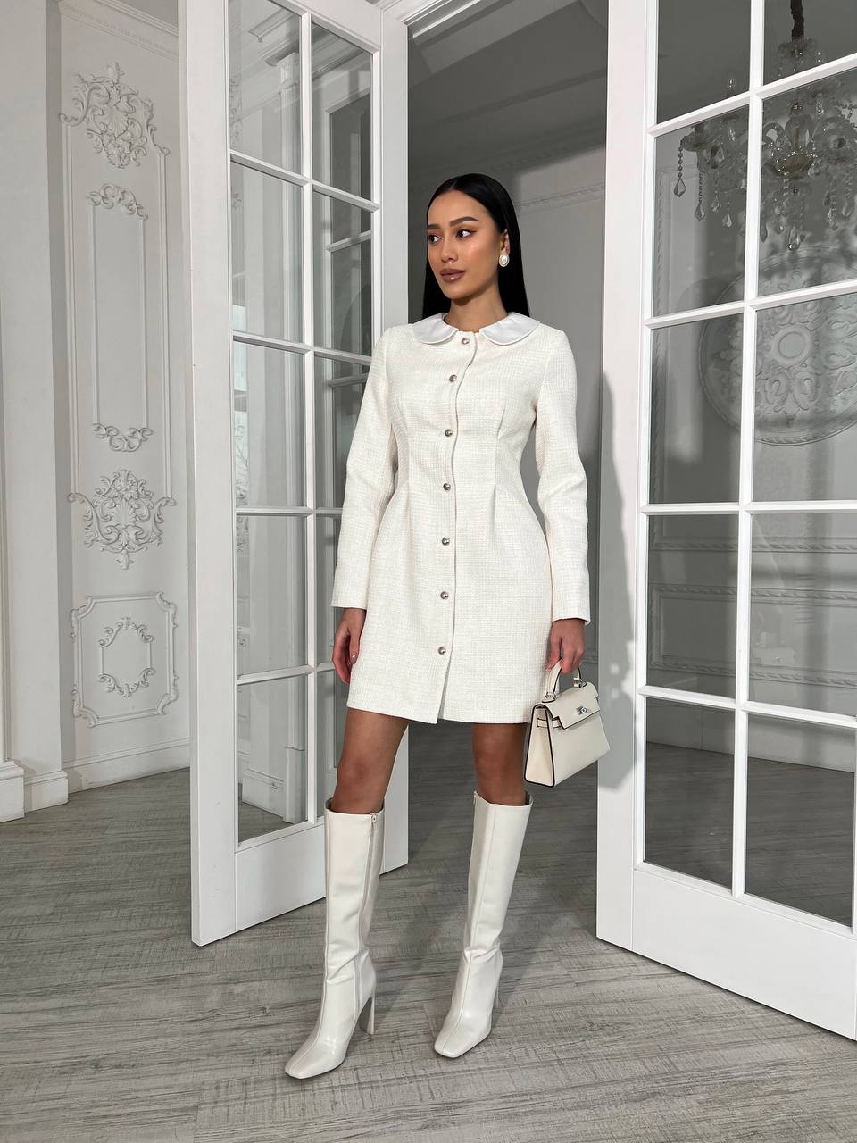 Woman in a white coat and knee-high boots standing in a room with large windows.