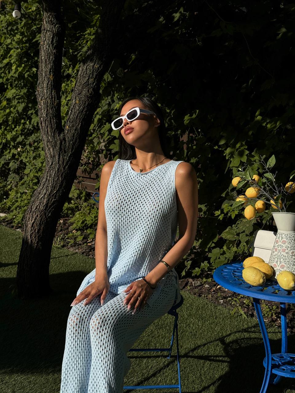 Woman in a light blue sleeveless top and pants sitting outdoors with sunglasses on, near a table with lemons.