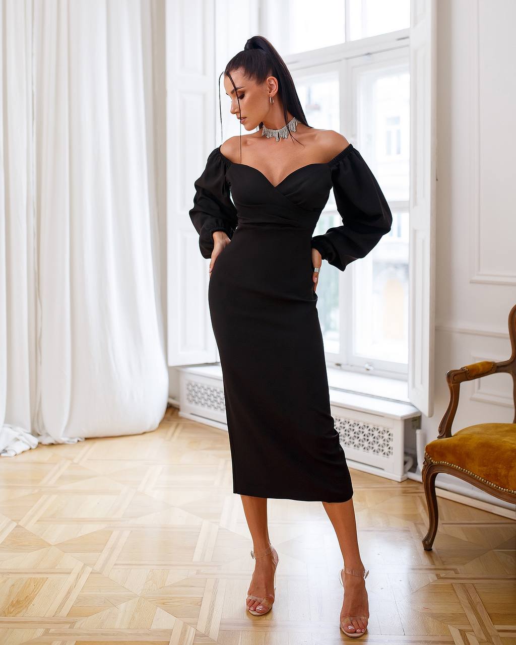 Woman wearing a black off-shoulder dress in an elegant room with white curtains and a wooden chair.