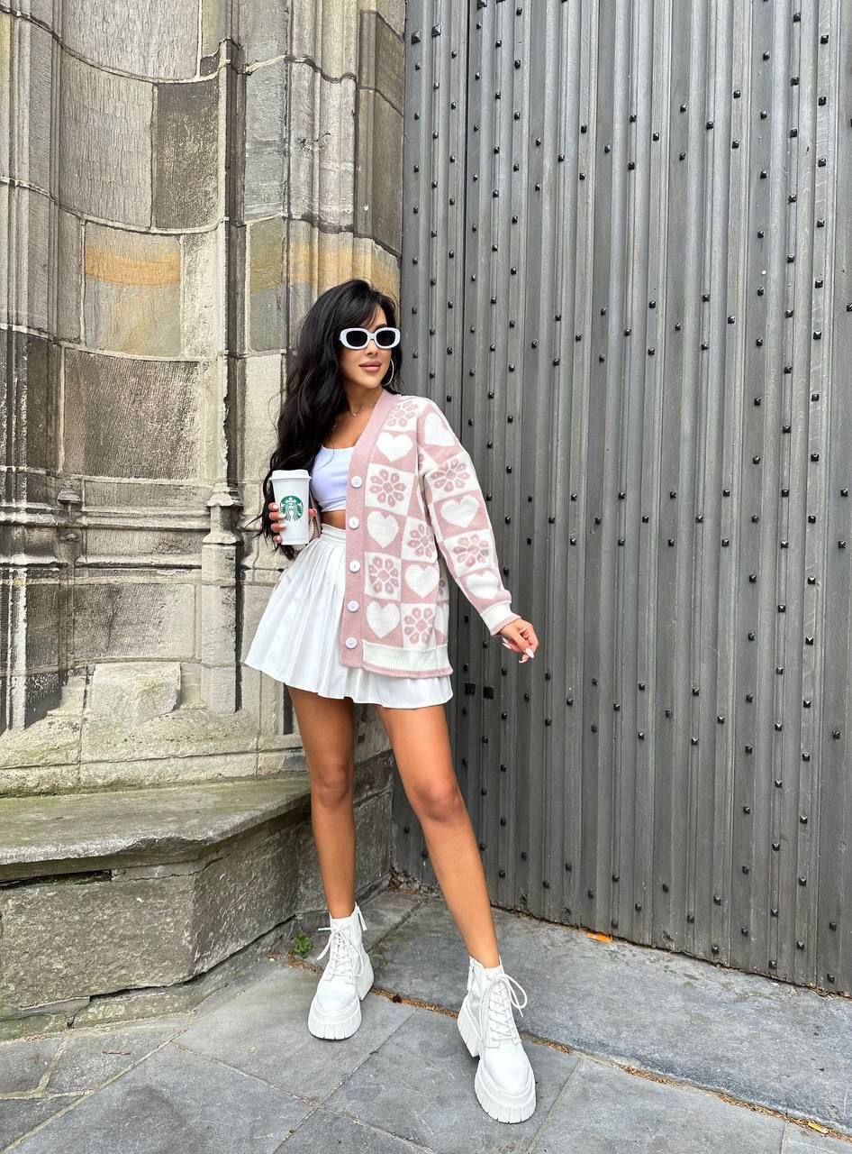 Woman in a white outfit with floral cardigan holding a coffee cup, standing against a stone wall.