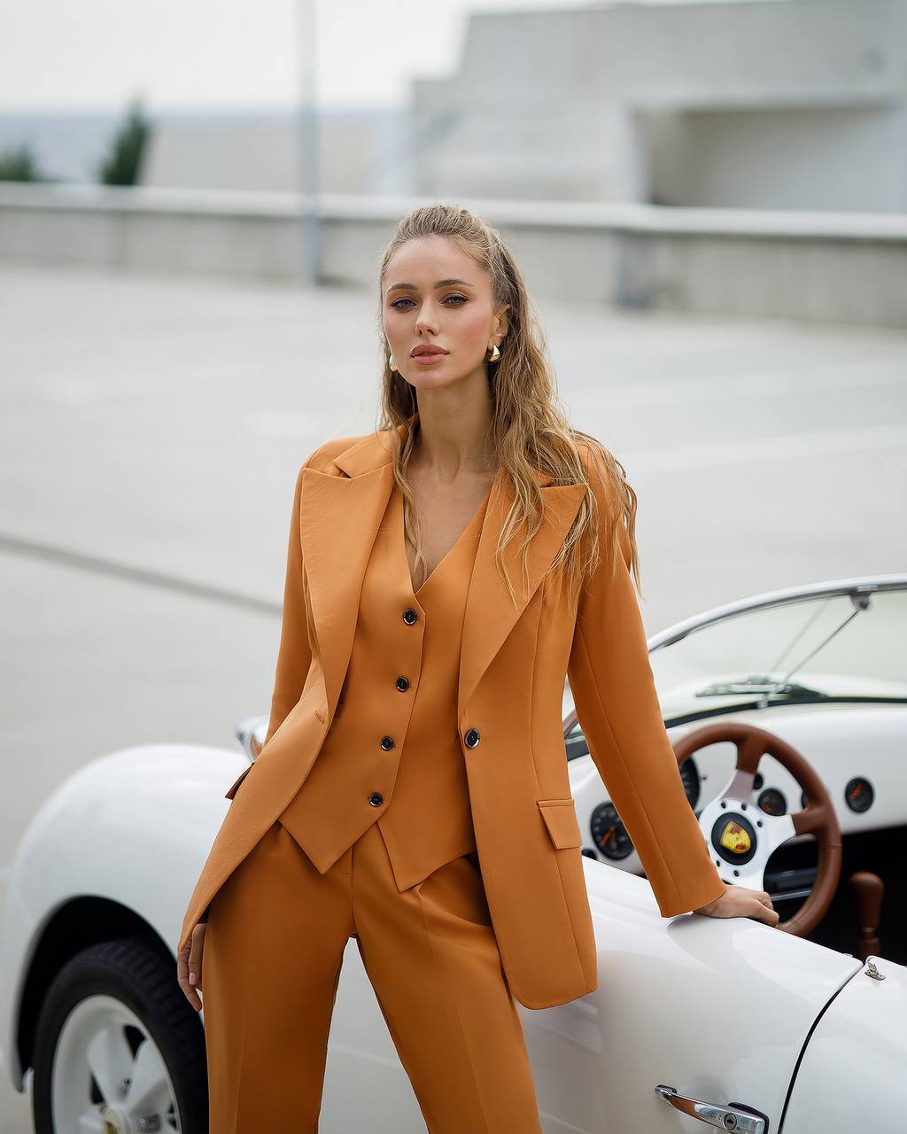 Woman in a mustard yellow suit standing next to a vintage car.