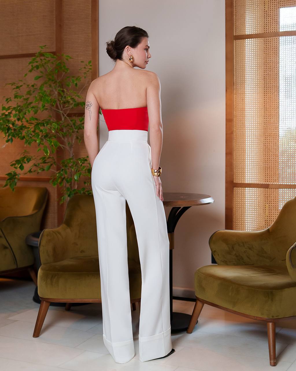 Woman wearing a red top and white pants in an indoor setting with chairs and a plant.