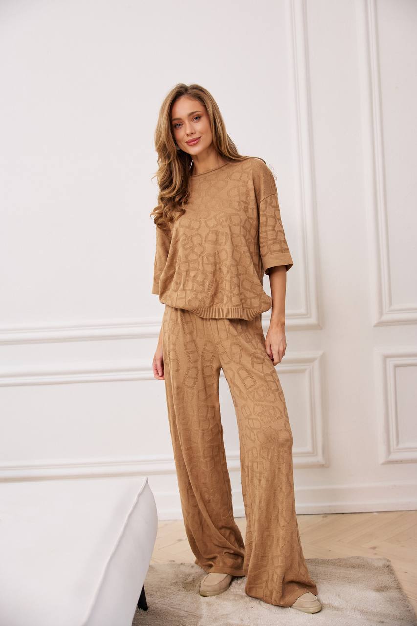 Woman wearing a brown textured outfit standing against a white wall.