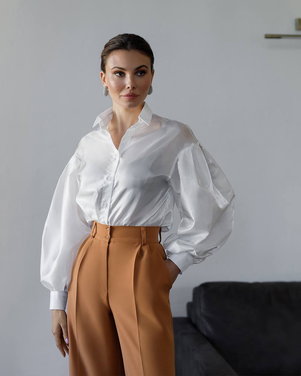 Woman wearing a white blouse with sheer sleeves and brown pants against a plain background