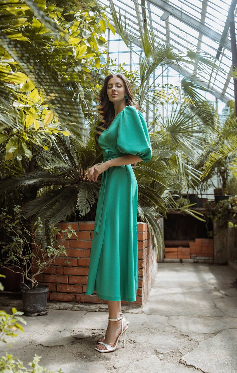 Woman in a green dress standing among plants in a greenhouse.