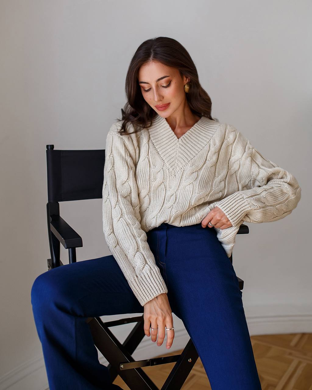Woman wearing a cream cable knit sweater and blue pants sitting on a chair.