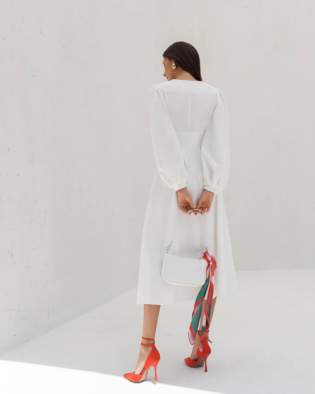 Woman in a white outfit with red shoes holding a white handbag against a white background