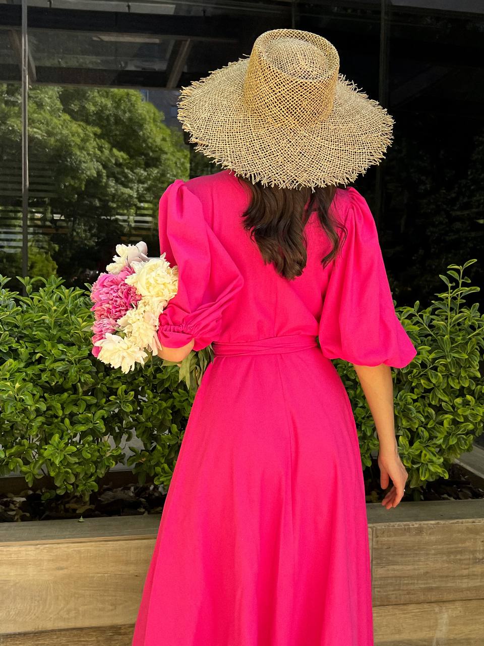 Woman in a bright pink dress and straw hat holding flowers, standing outdoors.