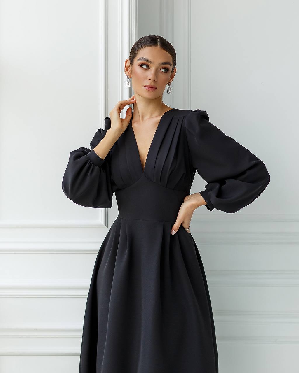 Woman wearing a black dress with puffed sleeves against a white wall.