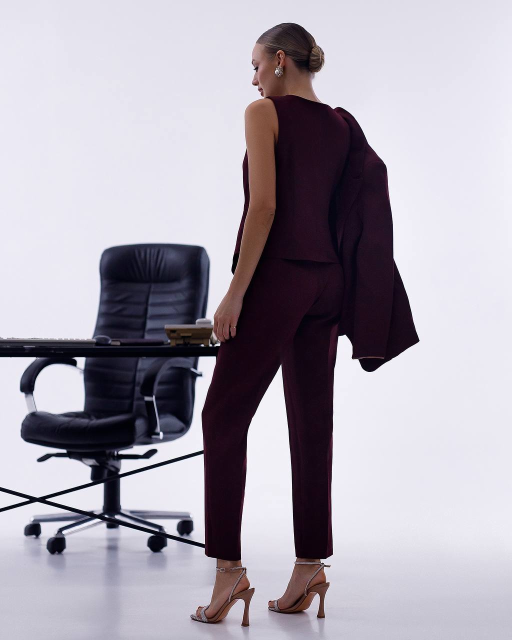 Woman in a dark suit standing in an office setting with a chair and desk.