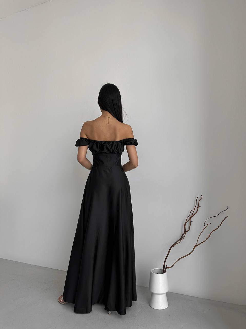 Woman wearing a black off-shoulder gown standing against a plain wall with a white vase and branches.