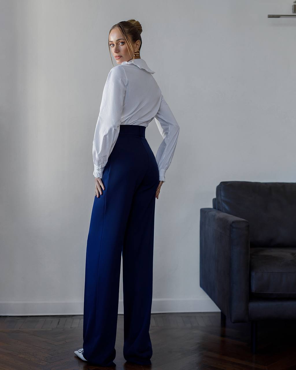 Woman wearing a white blouse and blue pants standing in a room with a gray wall and dark sofa.