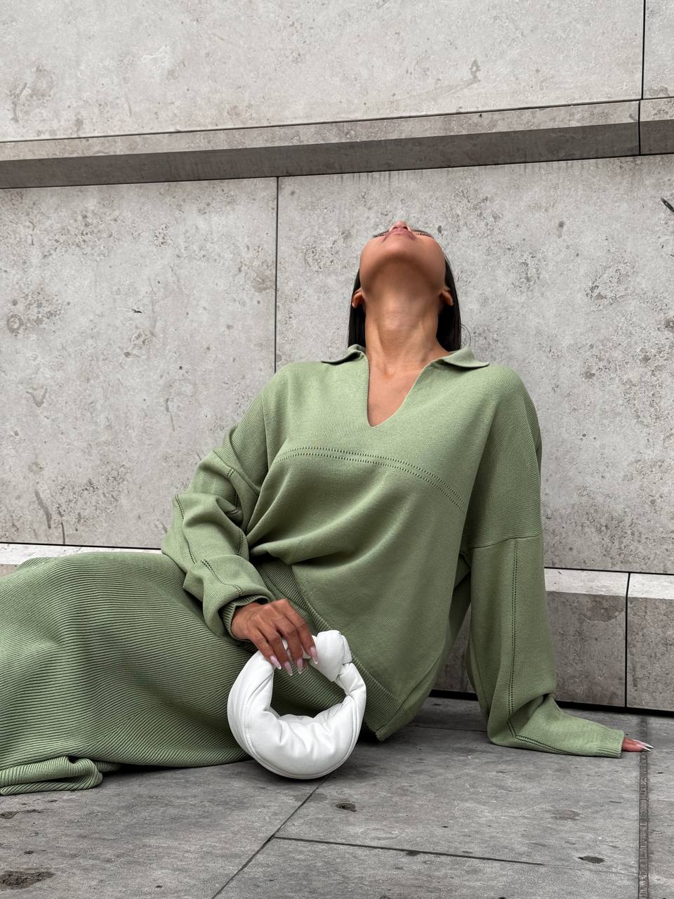 Person wearing a green outfit sitting on the ground with a white bag.