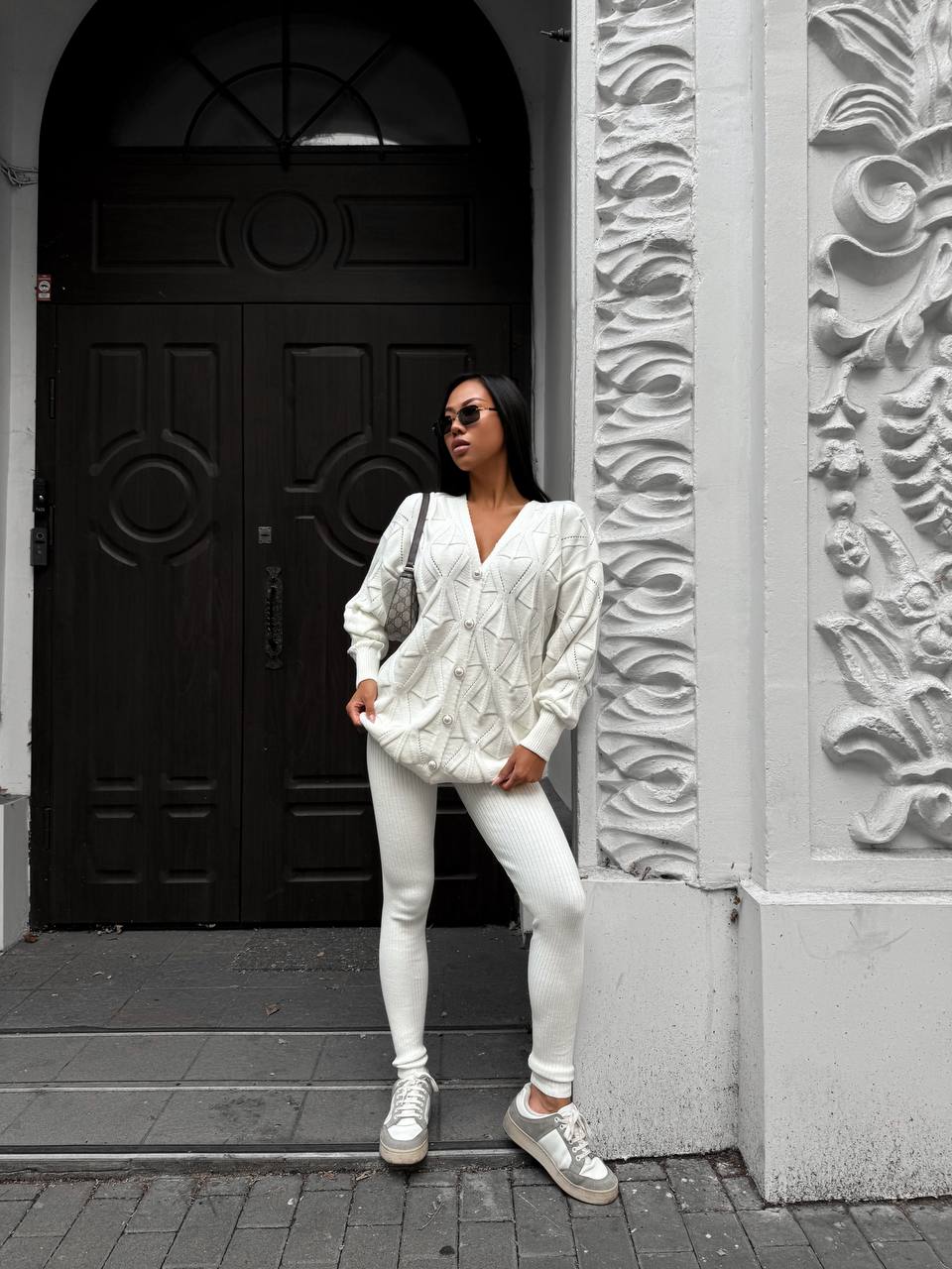 Woman in a white outfit standing in front of a black door with decorative patterns.