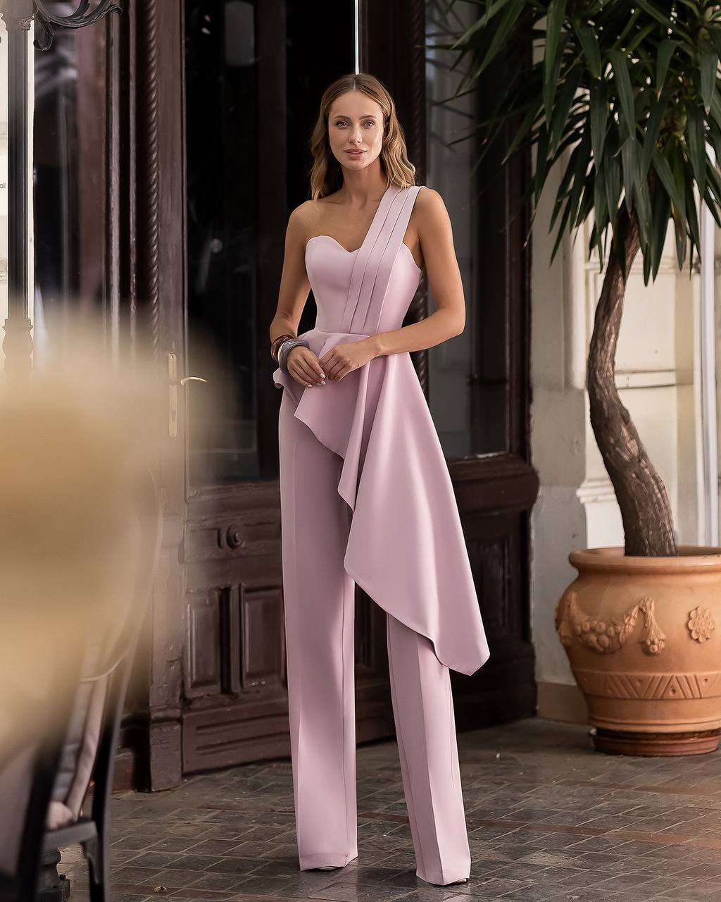 Woman wearing a pink one-shoulder jumpsuit standing indoors with decorative elements.