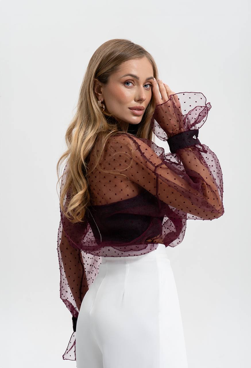 Woman wearing a sheer burgundy top with white pants on a light gray background