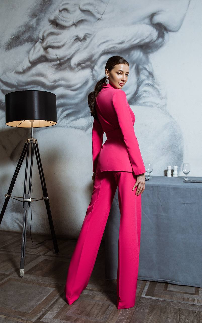 Woman in a bright pink suit standing in an elegant room with a lamp and statue.