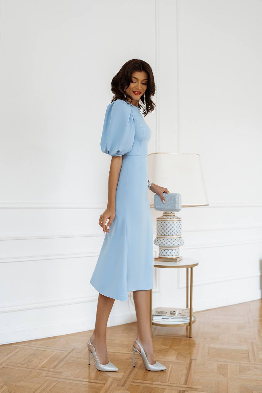 Woman in a light blue dress holding a clutch in a room with a white wall and wooden floor.
