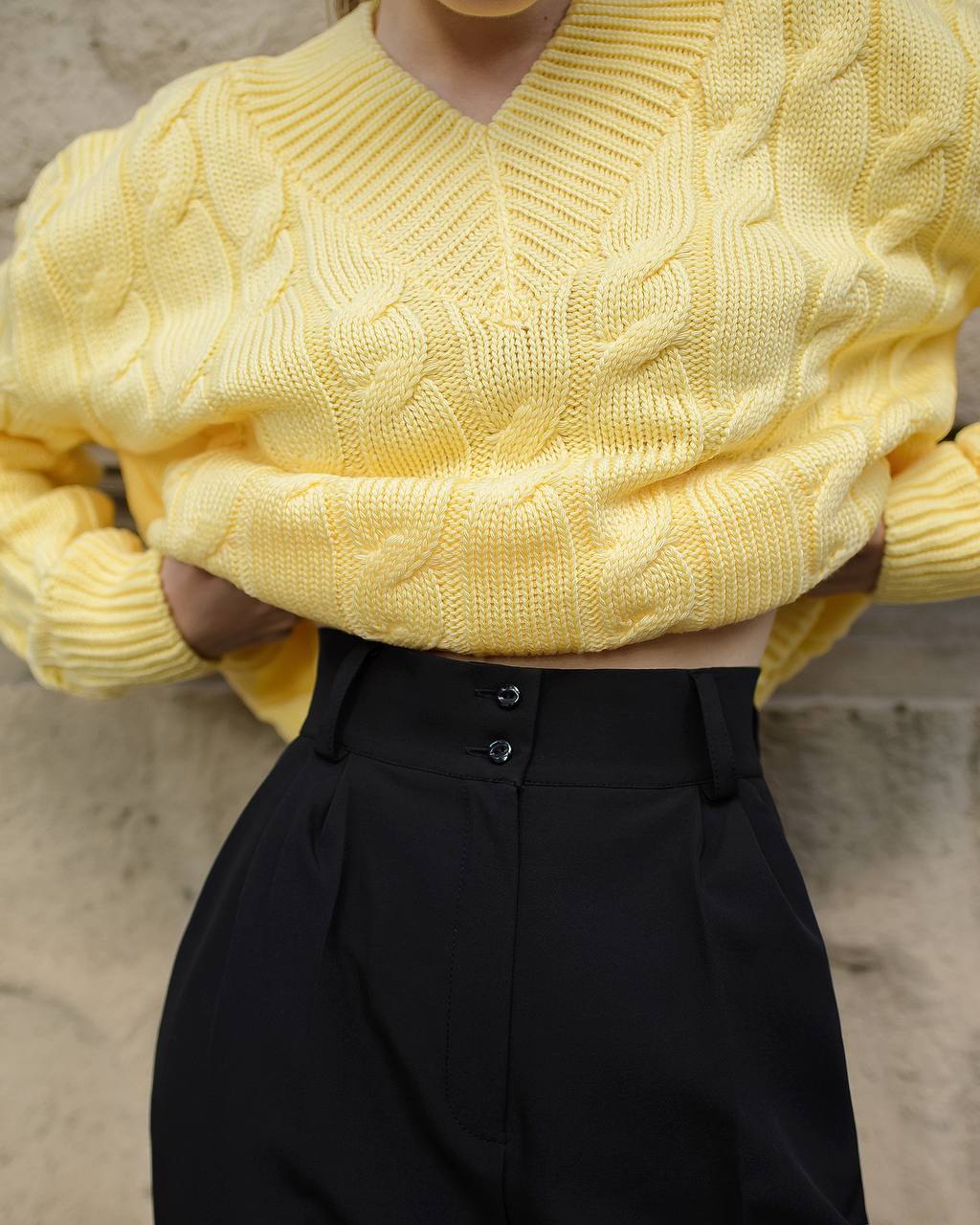 Person wearing a yellow cable knit sweater and black pants against a neutral background