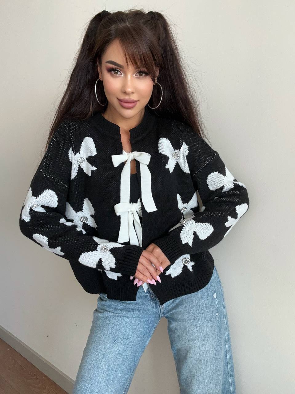 Woman wearing a black sweater with white bow patterns and light blue jeans against a plain background