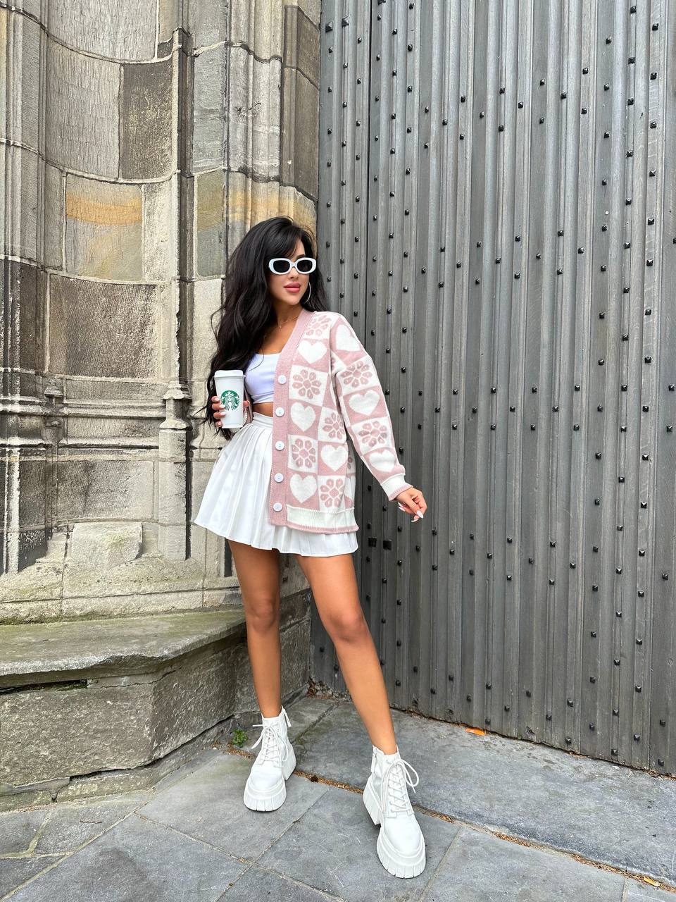 Woman in a white dress and pink cardigan standing in front of a stone wall.