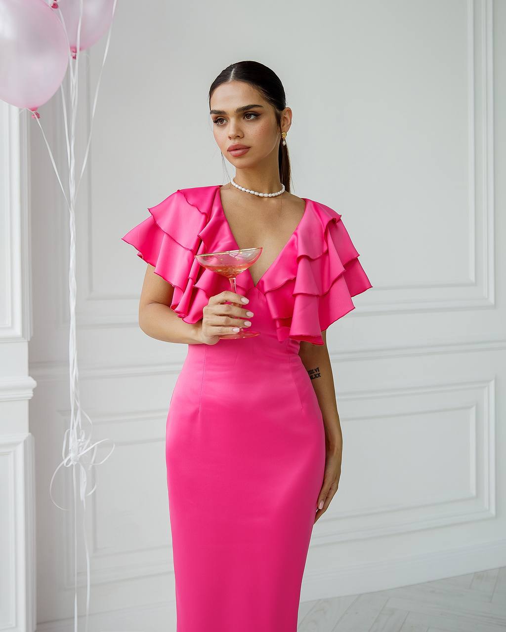 Woman in a bright pink dress holding a glass against a white wall with balloons.