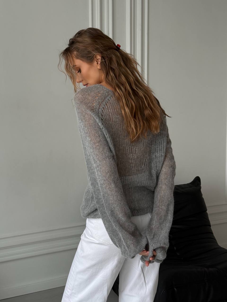 Woman wearing a gray sweater and white pants in a minimalistic room.