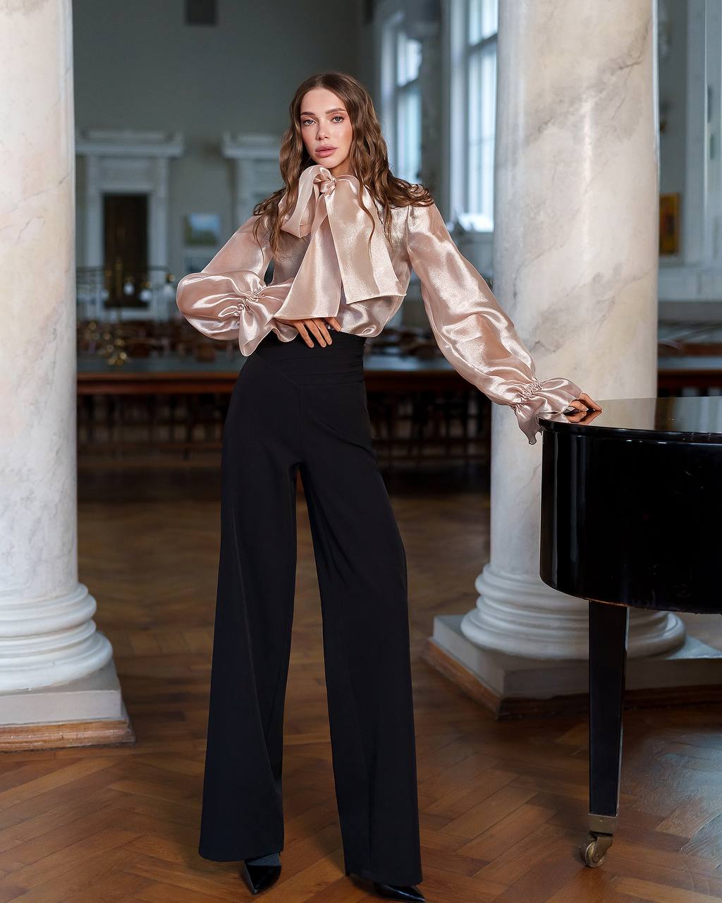 Woman in a satin blouse and black pants standing in an elegant interior setting.